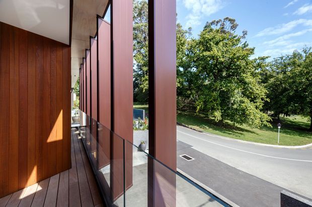 A narrow elevated exterior balcony or walkway beside a modern building, with warm vertical timber wall cladding on the left and a rhythmic series of tall rust-red privacy or sun-screen panels along the glazed edge. Timber decking, clear glass balustrading, and a landscaped suburban street with mature trees are visible beyond. Bright daylight and strong geometric shadows emphasize the contemporary facade.
