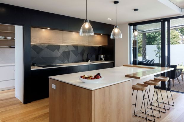Contemporary open-plan kitchen with a large light-wood island, white stone benchtop, dark cabinetry, warm timber flooring, and a geometric dark patterned splashback. Three glass pendant lights hang above the island, while large glazed doors open to an outdoor deck and bring in abundant natural light. A dining area is visible beyond the kitchen.