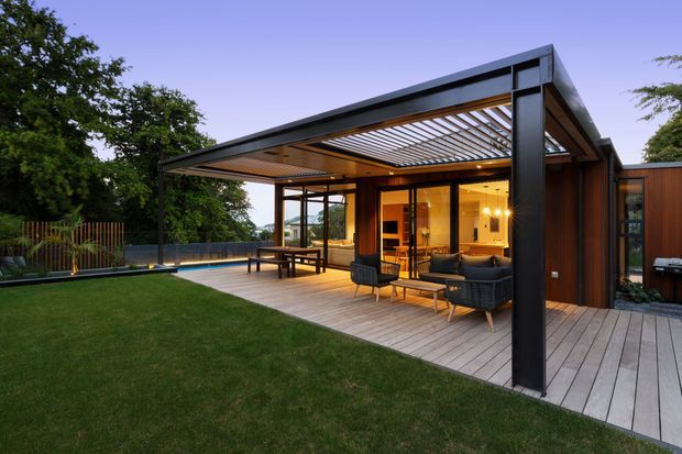Contemporary single-storey outdoor entertaining area with a flat-roofed pavilion, dark structural framing, warm timber soffit and decking, and extensive glazed walls opening onto a lawn. The covered terrace includes outdoor lounge seating and a dining table, with a partially enclosed interior living area visible behind the glazing. The setting is landscaped and leafy, photographed at dusk with warm interior lighting.
