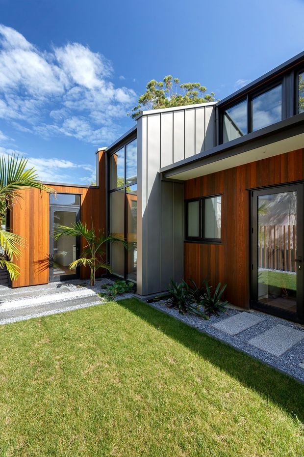 Exterior view of a contemporary multi-wing residential building arranged around a landscaped courtyard. The facade combines vertical warm timber cladding, dark metal-framed glazing, and a prominent light grey metal-clad upper volume. Gravel garden beds, palms, low planting, lawn, and stepping-stone paths surround the entrance and glazed doors under a bright blue sky.