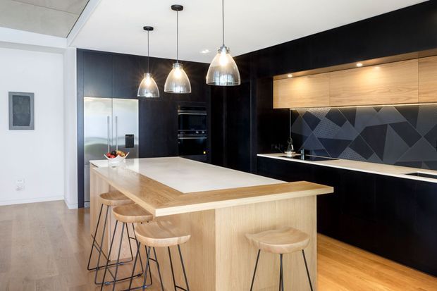Contemporary kitchen featuring a large light timber island with integrated seating, glossy black cabinetry, a geometric dark tiled splashback, timber flooring, and three pendant lights. The space is bright and open with a strong black-and-light-wood contrast.