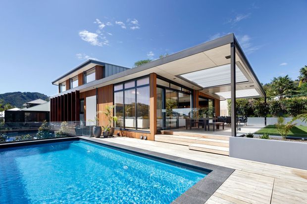 A contemporary two-storey residence with expansive floor-to-ceiling glazing, warm timber wall accents, and a broad flat-roofed outdoor living canopy. The home opens onto a pale timber pool deck beside a bright rectangular swimming pool, with landscaped planting and hills in the background.