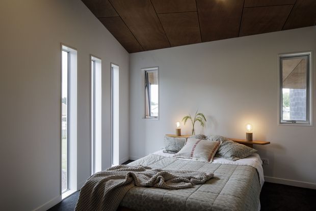 A calm, compact bedroom with a low-profile bed positioned beneath a dark timber-panelled ceiling. Pale grey walls, narrow vertical windows, soft neutral bedding, a simple timber headboard shelf, bedside candles, and a small plant create a warm, minimalist retreat. Dark carpet contrasts with the light walls and bedding.