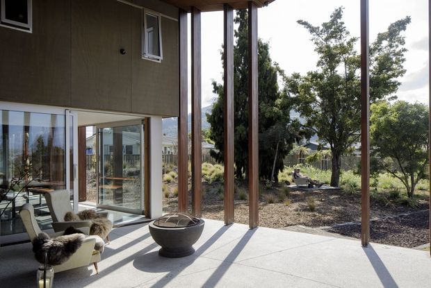 A modern covered outdoor terrace adjoining a glazed living area, with a concrete floor, slim timber posts, dark vertical exterior cladding, and large sliding glass doors. The terrace overlooks a landscaped garden and includes lounge seating and a central fire bowl. Strong sunlight creates long shadows across the spacious sheltered area.
