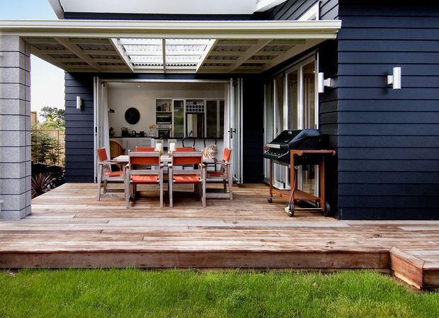 A covered outdoor dining and entertaining area opening from a contemporary home onto a timber deck. The space features a dining table with orange-accented chairs, large operable glazed doors, a built-in-style barbecue on a wheeled stand, dark horizontal exterior cladding, and a translucent roof panel above the pergola. Lawn is visible in the foreground.