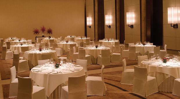 An elegant formal banquet or hotel ballroom arranged for a large seated dinner, with numerous round tables covered in white linens, place settings, floral centrepieces, slipcovered chairs, warm ambient wall lighting, and tall decorative wall panels.