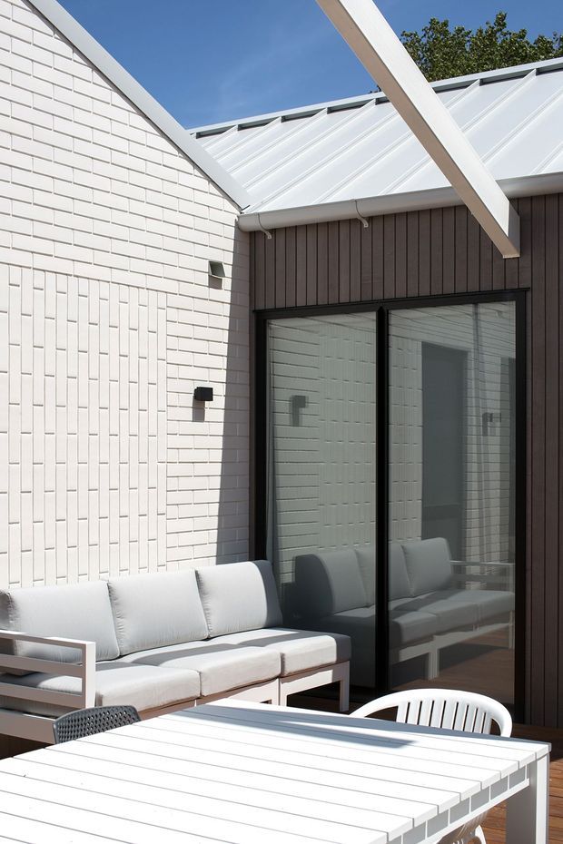 Bright contemporary courtyard or patio adjoining a building, with white brick walls, dark vertical cladding, black-framed glazed doors, and a white metal roof with exposed structural beams. A pale outdoor sofa and slatted dining table create a clean outdoor living area. Strong sunlight and crisp geometric lines give the space a refined, modern feel.