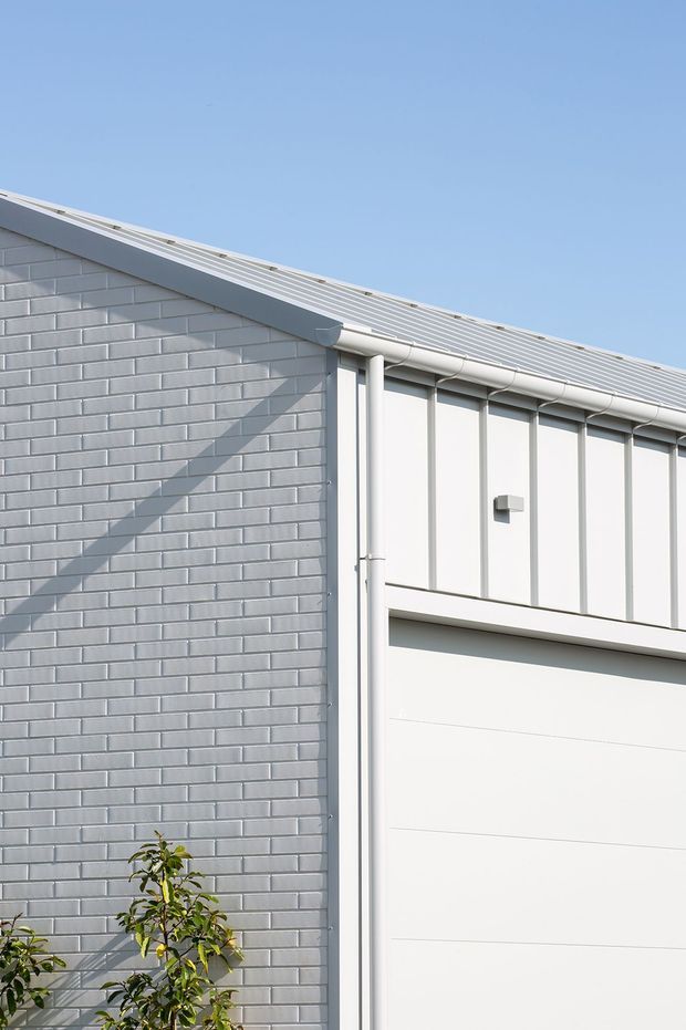 Close-up exterior view of a clean, light-coloured building facade. The image shows a grey brick gable wall adjoining a white vertically panelled wall and a large white sectional garage door. A light grey corrugated metal pitched roof with white gutters and downpipes runs across the top. Bright daylight, crisp shadows, and a small planted shrub appear at the lower edge.