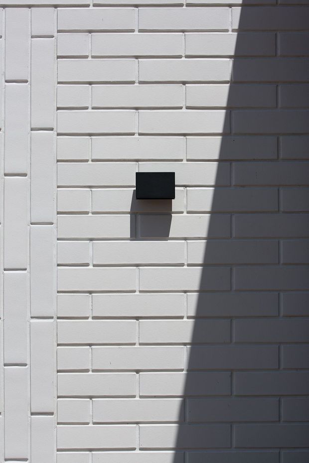 Close-up of a white-painted brick wall with regular horizontal masonry joints and a vertical brick detail at the left edge. A small black rectangular wall-mounted fixture projects from the wall, while strong sunlight and shadow create a sharp diagonal division across the surface.