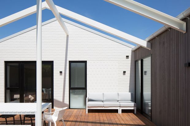 Bright exterior view of a contemporary courtyard-style residential terrace, with white horizontal cladding, dark vertical timber-like cladding, black-framed glazing, exposed white roof beams, and warm timber decking. Outdoor dining furniture and a sofa create a relaxed entertaining area under a clear blue sky.