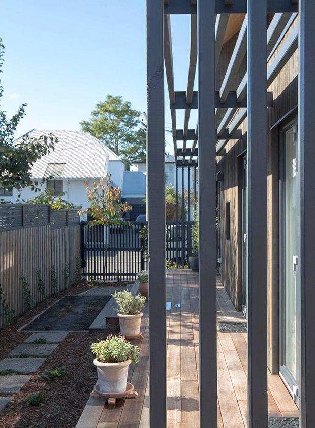 A narrow residential side courtyard and timber deck viewed along the side of a contemporary home. The deck is sheltered by a pergola with dark painted timber posts and beams, alongside a timber-clad wall and glazed doors. A landscaped garden, potted plants, slatted fencing, and neighbouring residential structures are visible beyond.