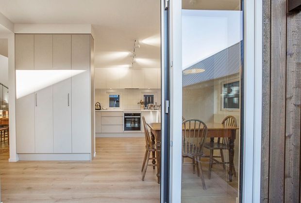 View through an open glazed door into a bright, modern kitchen with full-height white cabinetry, integrated appliances, timber flooring and a small dining area. The glazed opening leads to a covered courtyard or outdoor dining space with a timber table and chairs. A timber-clad exterior wall is visible at the right.