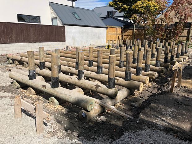 An outdoor construction site showing a partially assembled timber log structure made from large round treated posts and horizontal logs, with residential buildings and timber fencing in the background.