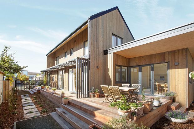 Exterior view of a contemporary timber-clad multi-level home with a steep gabled volume connected to a low-slung flat-roof wing. The house features extensive glazing, a timber deck with outdoor dining furniture and potted plants, a pergola-like entrance structure, and a landscaped path leading alongside the building.