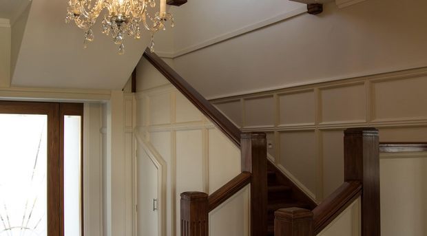 Interior view of a refined traditional stairway and entry area. The staircase has dark stained timber handrails, newel posts and balustrade details, cream painted panelled walls, decorative moulding, and a crystal chandelier. A timber-framed glazed entry door is visible at left.