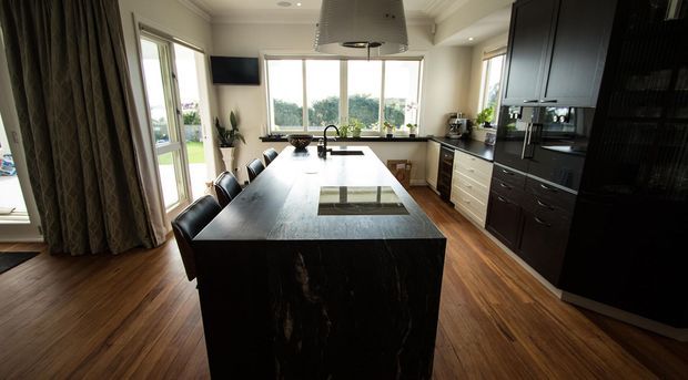 A spacious contemporary kitchen with a large dark stone island, integrated cooktop and seating, dark built-in cabinetry, pale benchtops, timber flooring, and abundant natural light from windows and glazed doors. A sculptural rangehood is suspended above the island, with a dining or living area visible beyond.