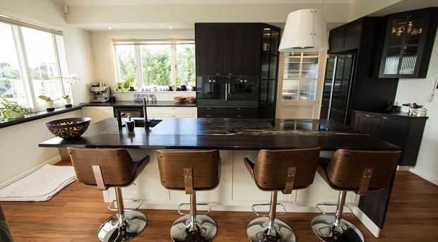 Bright contemporary kitchen with a large dark stone island and four timber bar stools, glossy dark cabinetry, integrated appliances, extensive windows, and warm timber flooring. A sculptural pendant light hangs above the island, while a white display cabinet and dark cabinetry add contrast.