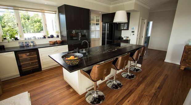 A spacious, refined kitchen with a large dark stone island, integrated appliances, white cabinetry, timber flooring, and four bar stools. Large windows bring in abundant natural light, while dark timber cabinetry and black accents add contrast.