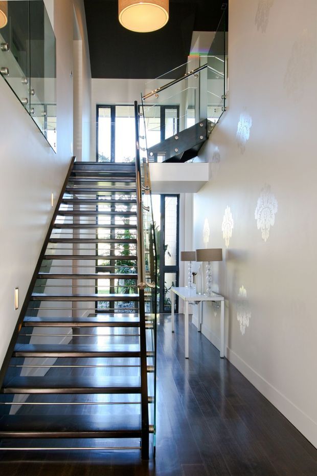 A bright, tall interior stair hall with an open-riser staircase, dark timber treads, black structural elements, and clear glass balustrades with metal handrails. The space features dark polished timber flooring, white walls with decorative patterned light projections, a high ceiling with a large pendant light, full-height glazing, and a small console with lamps along the right wall.