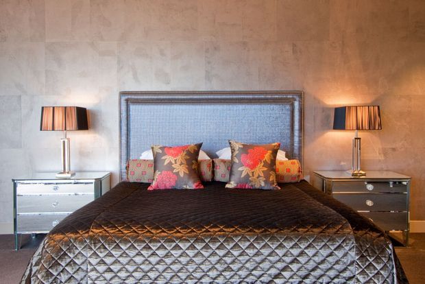 A moody, refined bedroom centered on a large bed with a quilted dark coverlet, upholstered textured headboard, and patterned floral accent cushions. Matching mirrored bedside chests and table lamps flank the bed, set against a warm textured plaster or concrete-look wall.