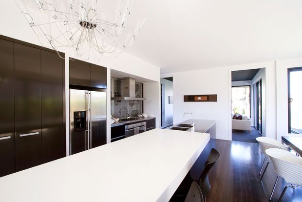 Bright contemporary open-plan kitchen with a large white island and integrated sink, dark full-height cabinetry, stainless-steel refrigerator and appliances, and dark timber flooring. A sculptural branching chandelier hangs above the island, with a dining area and living space visible beyond.