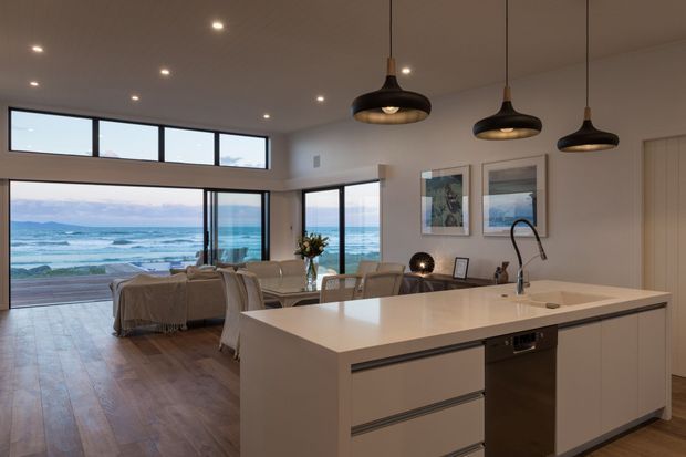 Contemporary open-plan kitchen and living area with a large white island, integrated sink and dishwasher, timber flooring, pendant lighting, and expansive glazing overlooking the ocean. A dining and lounge area sits beyond the kitchen, with framed artwork and a calm coastal outlook.