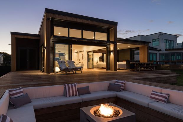 Contemporary two-storey home viewed at dusk from a landscaped outdoor entertaining area. The house features dark timber cladding, expansive floor-to-ceiling glazing, a broad timber deck, and an open living area connected to the exterior. In the foreground is a built-in upholstered outdoor lounge arranged around a square fire pit, with a dining table and chairs on the deck. A neighbouring modern home is visible to the right.