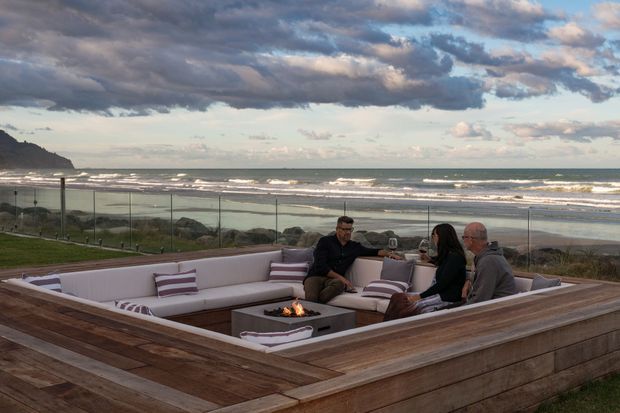An elevated outdoor lounge terrace overlooking a beach and ocean, furnished with a large built-in sectional seating arrangement around a central fire pit. Timber decking and a timber-clad raised enclosure frame the seating area, with glass wind screens and striped cushions. Several people are seated around the fire, beneath a dramatic cloudy coastal sky.