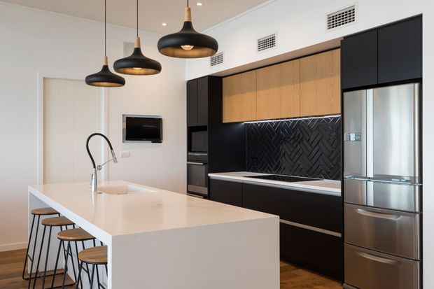Contemporary kitchen with a large white waterfall island and integrated sink, three black pendant lights, black and timber cabinetry, a dark herringbone tile splashback, stainless-steel refrigerator, and timber flooring. A small dining or living area is visible beyond the kitchen.
