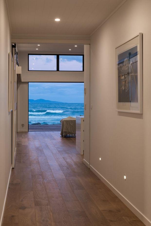 A warm, narrow interior hallway with wide plank timber flooring, light painted walls, and a painted paneled ceiling. Recessed floor-level and ceiling lighting illuminate the corridor, which opens toward a bedroom or living area with a covered bed or seating. Large windows and an open glazed door frame a clear ocean view with waves and distant mountains. A framed artwork hangs on the right wall, while built-in cabinetry or shelving is visible along the left.