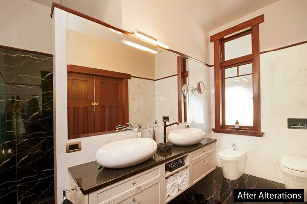 Elegant renovated bathroom with a double floating-style vanity, two white vessel basins, dark stone benchtop, white marble-look wall tiles, dark marble-look floor and shower wall, warm timber cabinetry, and leaded-style timber-framed windows. A large illuminated mirror spans the vanity, with a separate toilet visible at right.