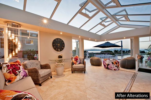 Bright, spacious indoor-outdoor living area with a glazed roof featuring multiple operable skylights and extensive openings to a waterfront terrace. The room includes upholstered lounge seating, pendant lighting, a central coffee table, tiled flooring with decorative border detailing, and a large wall clock. An outdoor umbrella, water view, and landscaped surroundings are visible beyond the open glazing.