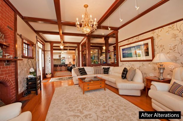 A warm, traditionally styled living room with polished timber flooring, exposed dark timber ceiling beams, patterned wallpaper, and a red brick fireplace. The room features cream upholstered sofas, a carved timber coffee table, a large patterned rug, a crystal chandelier, framed artwork, and timber-framed glazing. A dining or kitchen area is visible beyond the living space.