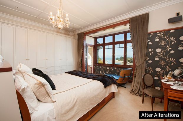 Elegant traditional bedroom with a large upholstered bed, built-in white wardrobes, ornate chandelier, patterned floral wallpaper, rich timber-framed bay window, and full-length drapery. The room has refined period detailing and a warm, classic palette, with a small desk and seating area near the window.