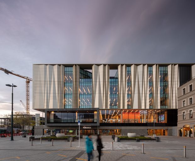 Exterior view of a large contemporary commercial building in an urban paved plaza. The facade combines tall vertical pale panels with extensive blue-grey glazing, a dark recessed lower volume, and a brightly illuminated orange-toned interior at ground level. Street furniture, bollards, pedestrians, adjacent buildings, and a construction crane are visible around the building.