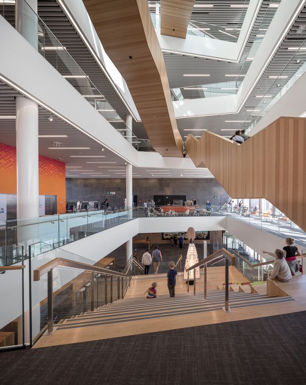A large, bright multi-level public interior with broad staircases, glass balustrades, open balconies, white structural columns, timber-lined ceiling and wall elements, and extensive linear ceiling lighting. People occupy the circulation spaces and a café or reception area is visible across the atrium. The architecture is contemporary and spacious, with strong visual connections between multiple floors.
