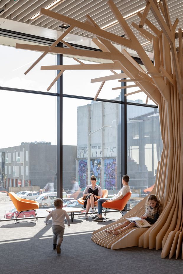 A bright contemporary public interior or commercial lounge with floor-to-ceiling windows overlooking an urban streetscape. The space features a dramatic sculptural timber installation resembling branching tree trunks and limbs, integrated with built-in seating along the right wall. Orange mid-century-style lounge chairs, small round tables, and people using the seating occupy the carpeted area. The ceiling has exposed linear white slats with recessed lighting, while dark-framed glazing creates a strong connection to the city outside.