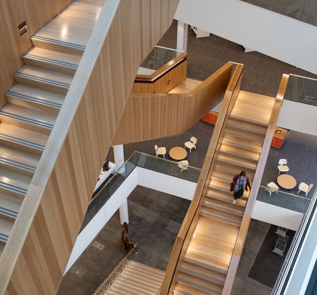 A high-angle interior view of a large contemporary multi-level atrium dominated by several timber-lined staircases and landings. The stairs feature light timber treads, slim metal or glass balustrades, and integrated linear lighting. White walls, dark tiled flooring, glass edges, and small round café-style tables with chairs are visible across the open circulation space. People are present on the stairs and lower floor. The composition emphasizes the complex vertical connections and open stairway arrangement.