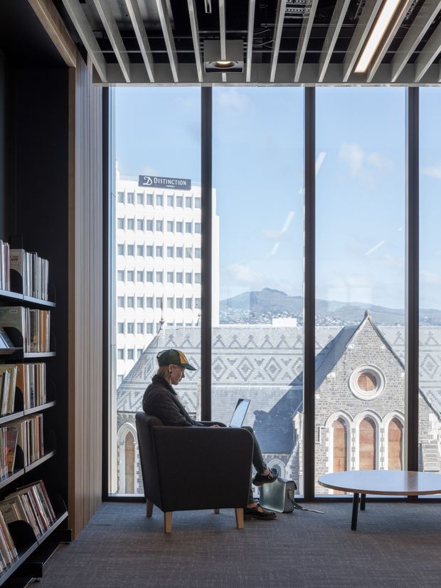 A contemporary commercial interior lounge or reading area with floor-to-ceiling windows overlooking a city building, a historic stone church roofline, and mountains. A person sits in a dark armchair using a laptop beside a small round table. Built-in dark shelving filled with books lines the left wall. The ceiling features exposed linear beams, integrated lighting, and visible services, while the floor is carpeted in dark grey.
