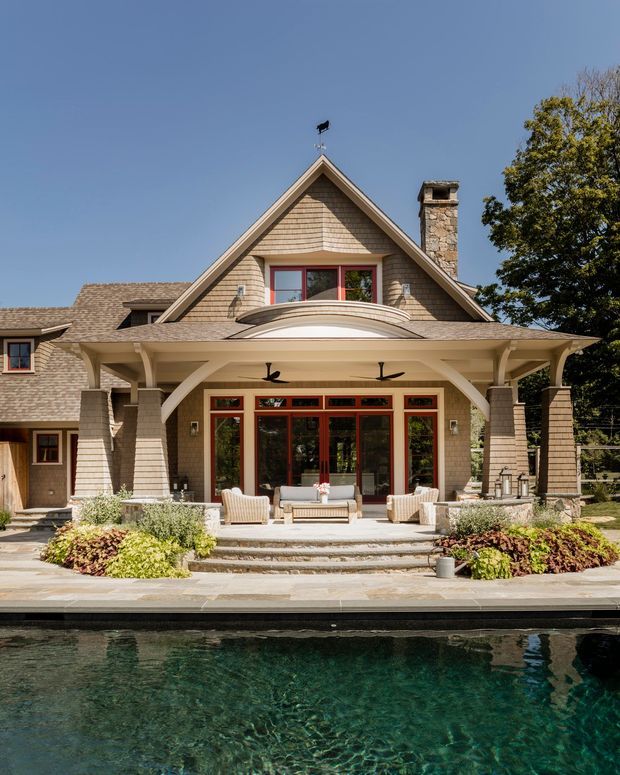 A substantial shingle-clad house viewed from across a swimming pool. The façade features a steep front-facing gable, broad covered patio, curved timber or painted arches, red-framed glazing and doors, a stone chimney, and a projecting curved balcony. Outdoor seating and landscaped planting occupy the terrace.