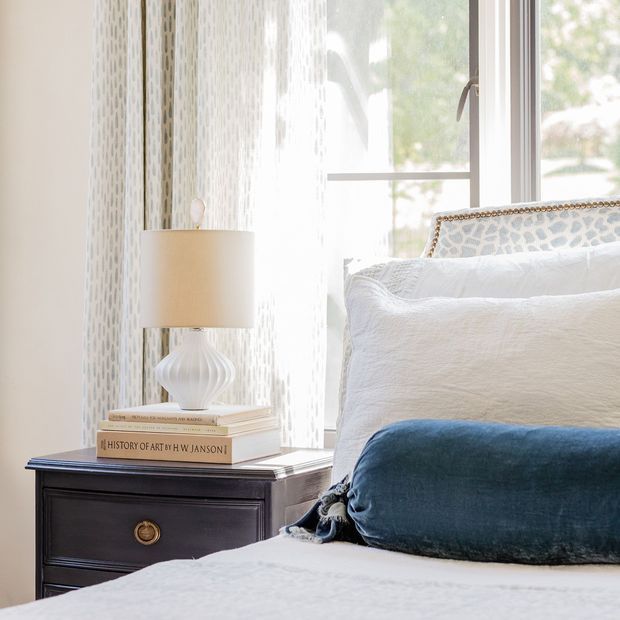 Bright, refined bedroom vignette featuring a dark wood bedside table with brass drawer pull, a white ceramic table lamp, stacked books, textured white bedding, and a deep blue velvet accent cushion. A large window with sheer patterned curtains provides abundant natural light, while a decorative upholstered headboard adds a classic touch.
