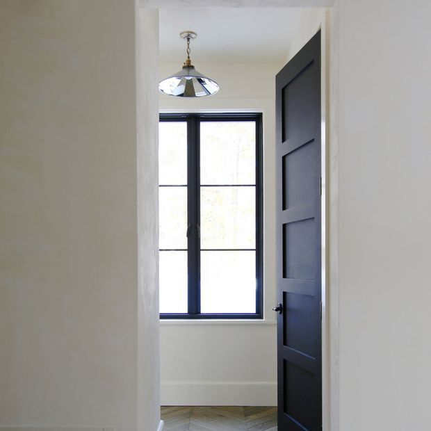 A compact, bright interior viewed through an open doorway, featuring a dark navy or charcoal panelled door, black-framed multi-pane window, white walls and trim, and a small pendant light with a metallic reflective shade. A herringbone-patterned floor is visible at the threshold and beneath the window.
