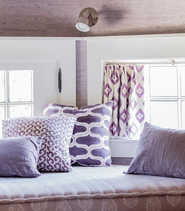 Bright close-up view of a styled bedroom with a bed layered in lavender and white patterned pillows and bedding. White wall panelling, a wood-look ceiling, divided windows, and coordinating geometric curtains create a fresh, decorative interior.