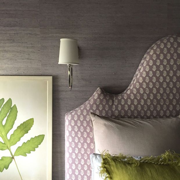 Close-up view of a refined bedroom corner featuring a tall upholstered bedhead in patterned mauve fabric, layered neutral and olive cushions, a wall-mounted bedside lamp, dark textured wall cladding, and framed botanical artwork.