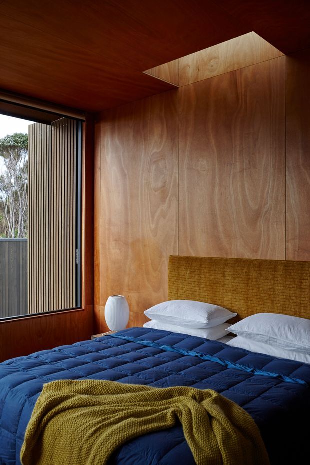 A warmly lit bedroom with extensive natural timber wall and ceiling panelling, a large window with vertical timber screening, and a bed dressed in deep blue bedding with a mustard knitted throw. A simple white bedside lamp sits beside the bed. A small angled ceiling opening or skylight brings additional light into the timber-lined room.