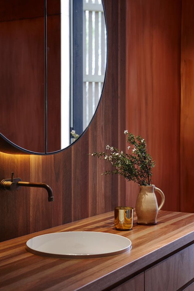 Close-up view of a sophisticated bathroom vanity with a round vessel basin set into a richly grained timber countertop. The walls and vanity cabinetry are clad in warm dark timber, with a slim dark metal wall-mounted tap and a large organically curved black-framed mirror. Soft concealed lighting beneath the mirror adds warmth, while a ceramic vase with branches and an amber glass add subtle decoration.