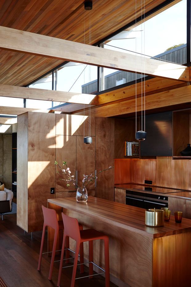 A warm, contemporary open-plan kitchen and dining area with extensive timber joinery, a large timber island with bar seating, integrated appliances, and dramatic exposed timber ceiling beams. Floor-to-ceiling glazing and clerestory windows bring in abundant natural light, while multiple black pendant lights hang over the island. A living area is partially visible beyond the kitchen.