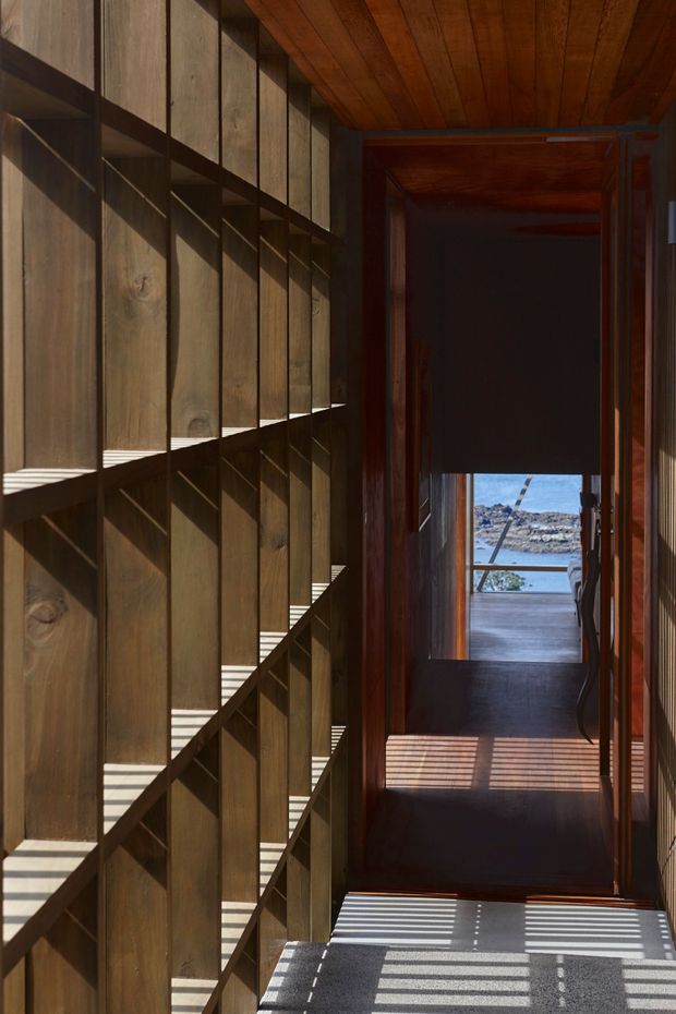 A narrow, dark timber-lined interior passage with strong sunlight casting striped shadows through a slatted or screened wall on the left. The ceiling, wall framing, doorway and flooring are all richly stained wood. The corridor leads toward an exterior deck or balcony, beyond which a body of water and rocky shoreline are visible. The image emphasizes materiality, geometric shadow patterns and a dramatic connection to the landscape.