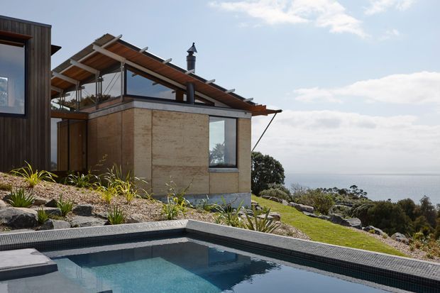 Contemporary hillside residence overlooking the ocean, with a flat-roofed masonry volume, extensive glazing, a timber-clad sloping roof structure, and a swimming pool in the foreground. The landscaped site uses exposed rocks and drought-tolerant planting, creating a strong connection between the house, pool, and coastal setting.