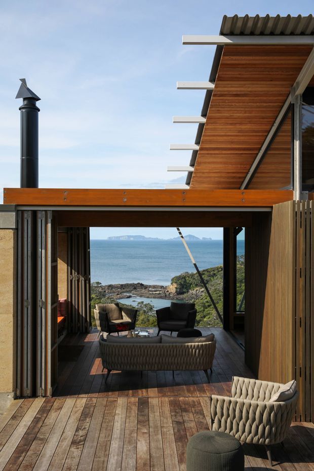 A contemporary coastal home with a covered timber deck opening toward expansive ocean views and rocky shoreline. The image highlights warm timber decking and soffit lining, dark structural framing, exposed roof projections, a corrugated metal roof, and an outdoor lounge furnished with woven chairs, a sofa, and an ottoman. A black cylindrical flue is visible at the left, while vertical timber screening frames the right side.
